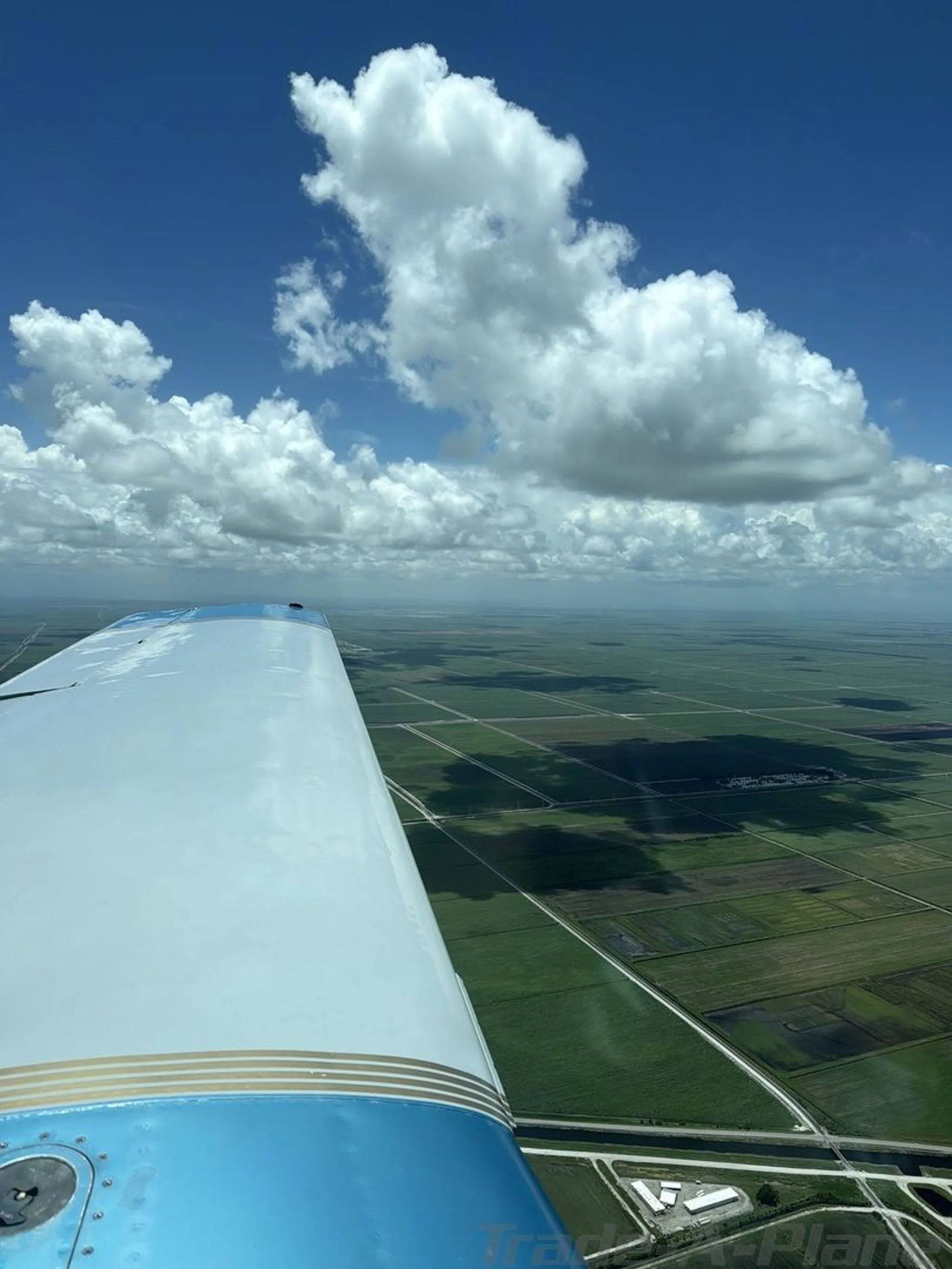 View from the wing of a light blue and white aircraft flying over a grid of green fields and waterways.