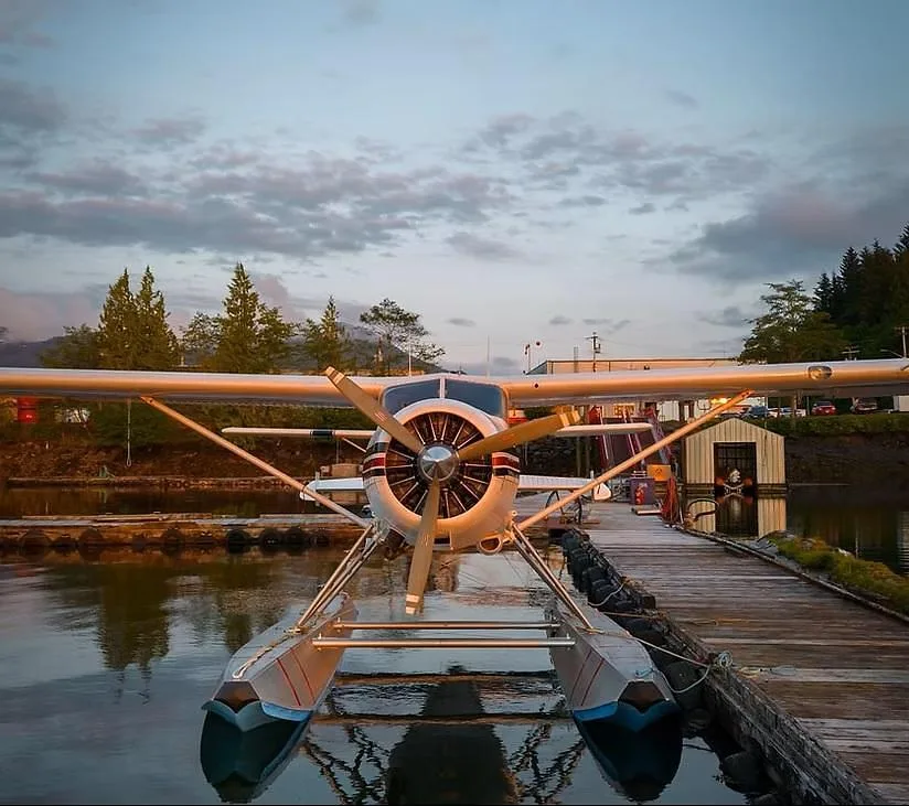 A white De Havilland Beaver seaplane with blue floats is docked at a wooden pier at dusk, with trees and buildings in the background.