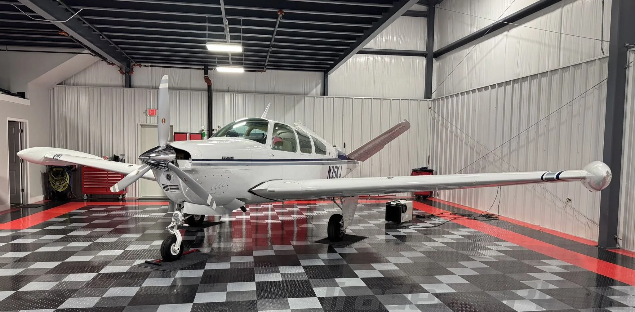 A front-side view of a white Beechcraft Bonanza with blue and grey stripes parked in a hangar.