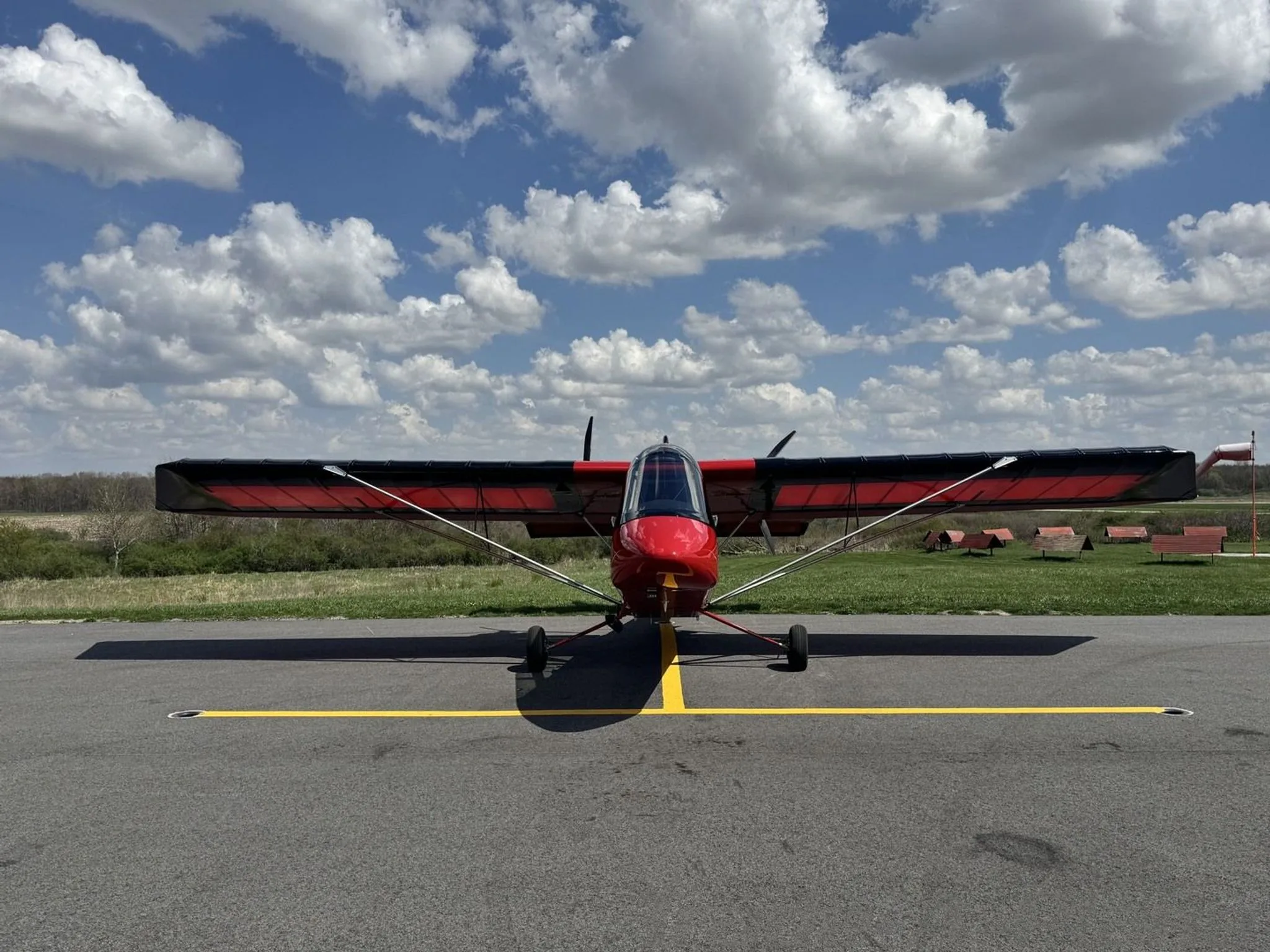 A red and black Air-Cam aircraft with twin engines is parked head-on on an airfield under a blue sky with white clouds.