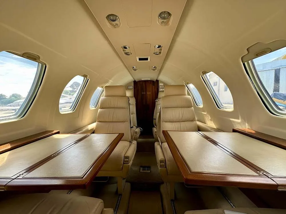 The aircraft cabin interior features light tan leather seats and two wooden tables, viewed looking towards the rear.