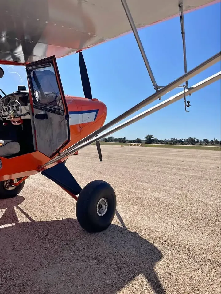 A low-angle view of an orange and silver high-wing aircraft with its door open on a gravel runway.