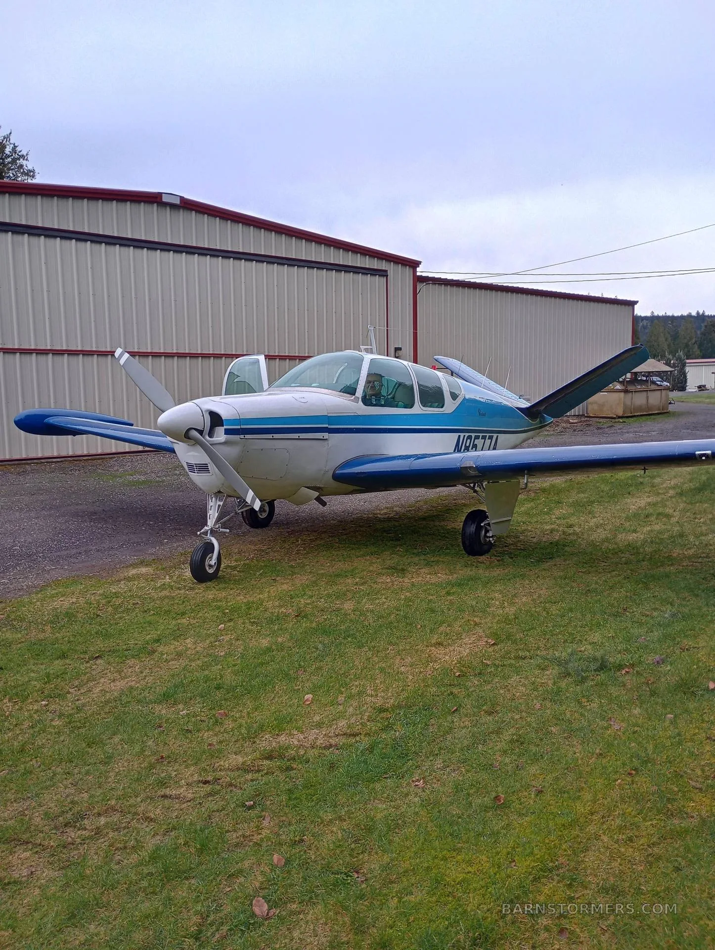 A white and blue single-engine aircraft, N1877A, parked on grass next to hangars under a cloudy sky.