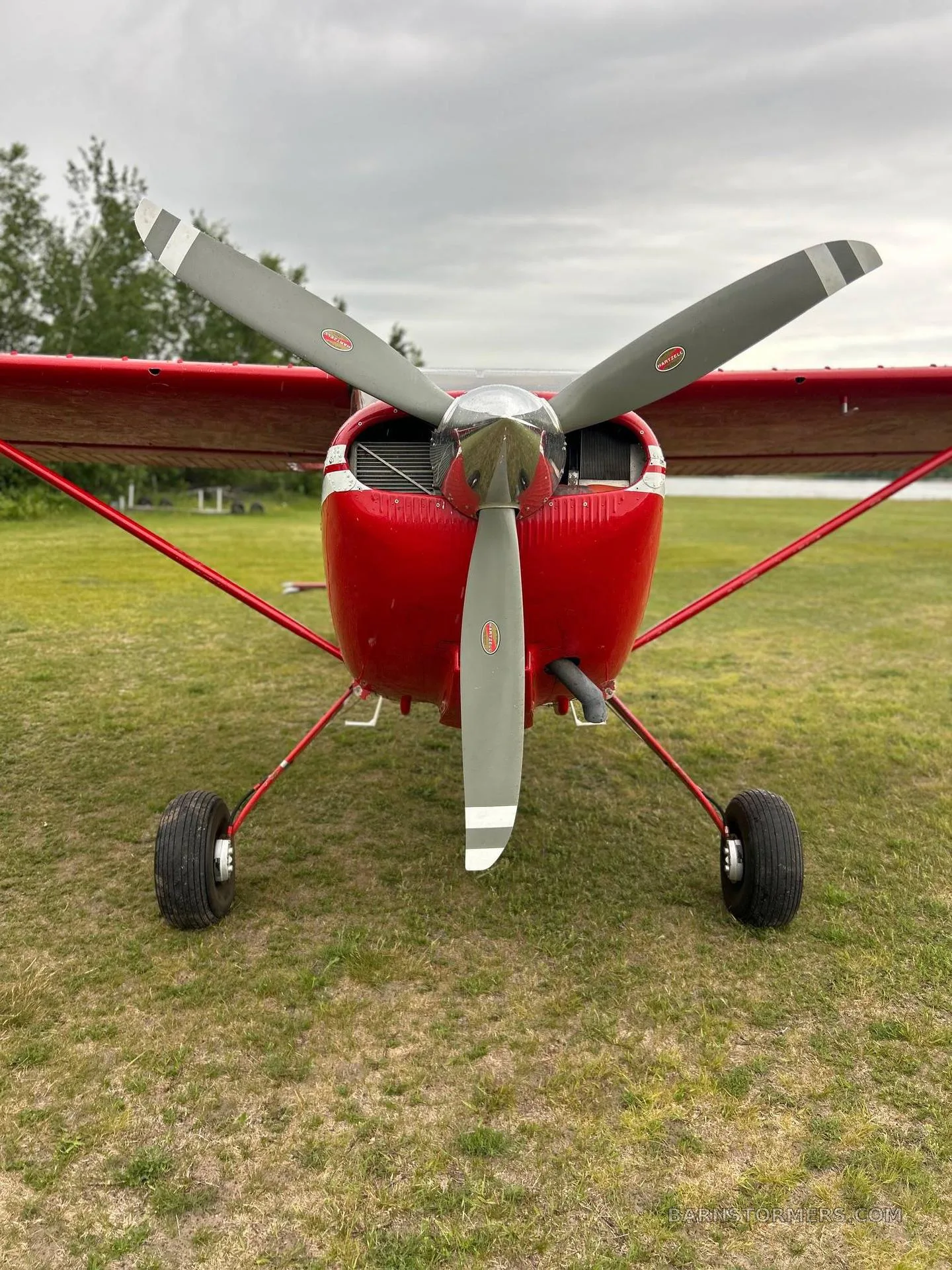 A front view of a red single-engine aircraft with a two-bladed propeller on a grassy field.