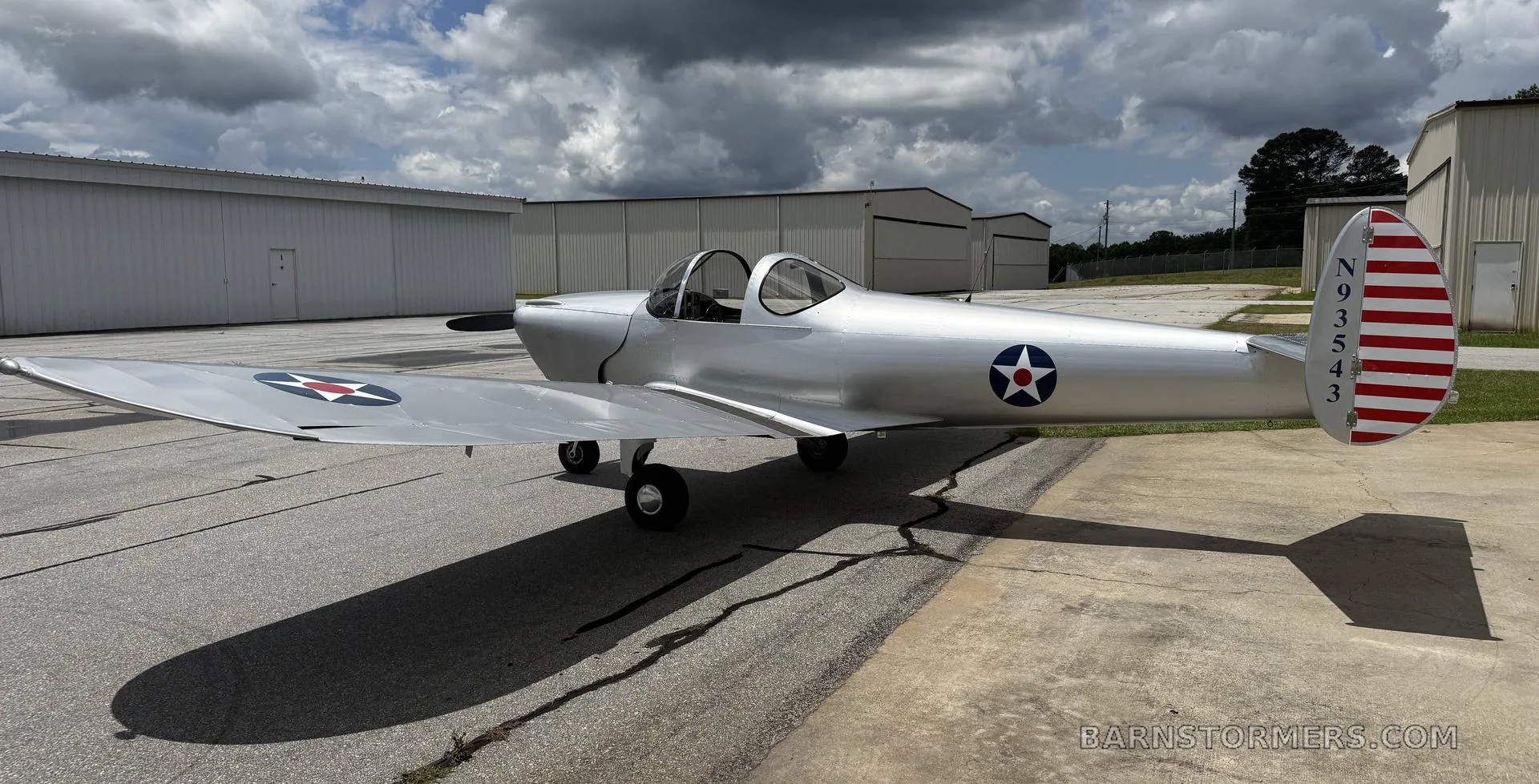 A silver single-engine aircraft with a bubble canopy and US military-style markings on the wings and tail, parked on an asphalt ramp.