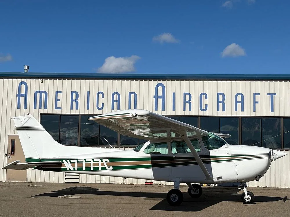 A side view of a white Cessna 172 with green and brown stripes, registration N7777C, parked on an airfield.