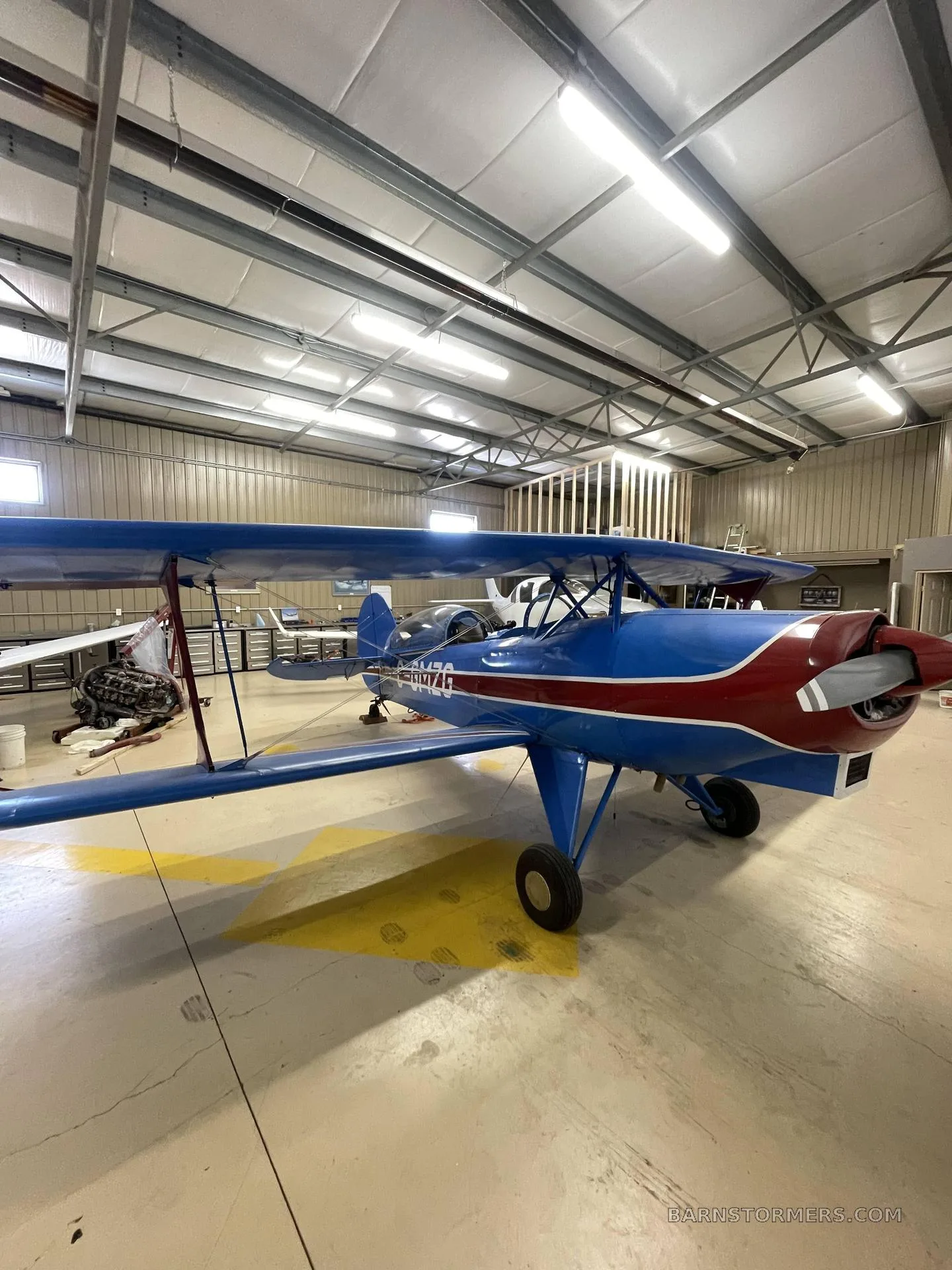 A blue and red biplane with registration C-GMZG is parked inside a brightly lit hangar.