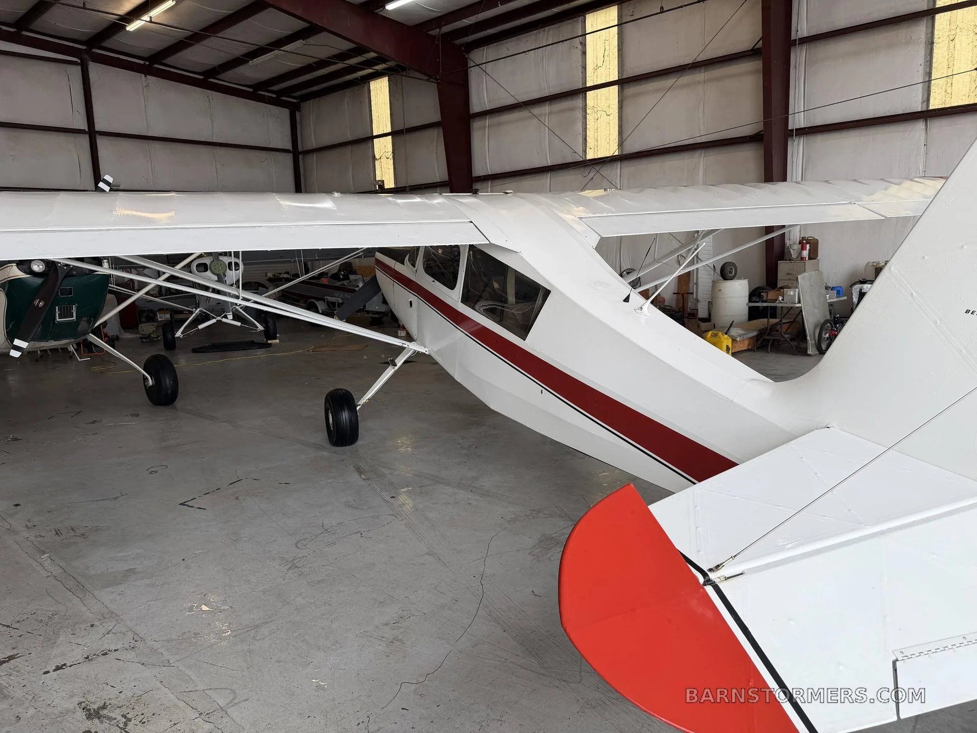 A rear-side view of a white and red high-wing aircraft parked in a hangar, showing the tail and right wing.