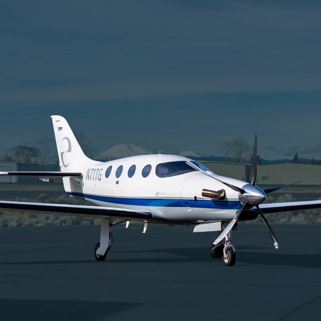 A white Epic E1000 GX aircraft with blue and grey stripes is parked on an airfield with mountains in the background.