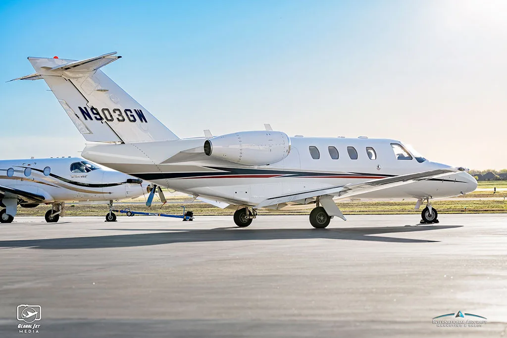 A side view of a white Cessna Citation CJ3 (N903GW) with blue and red stripes parked on an airfield.