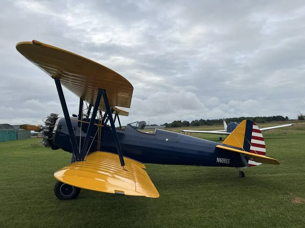 A blue and yellow biplane with a radial engine is parked on a grassy field under a cloudy sky, showing the N60955 registration on the tail.
