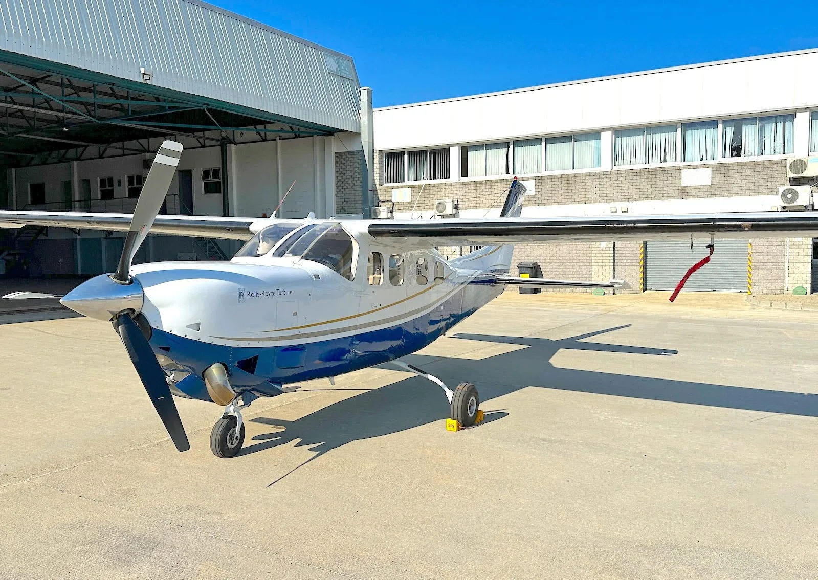 A white and blue single-engine turboprop aircraft, with 'Rolls-Royce Turbine' on its nose, parked on an airfield in front of a hangar.