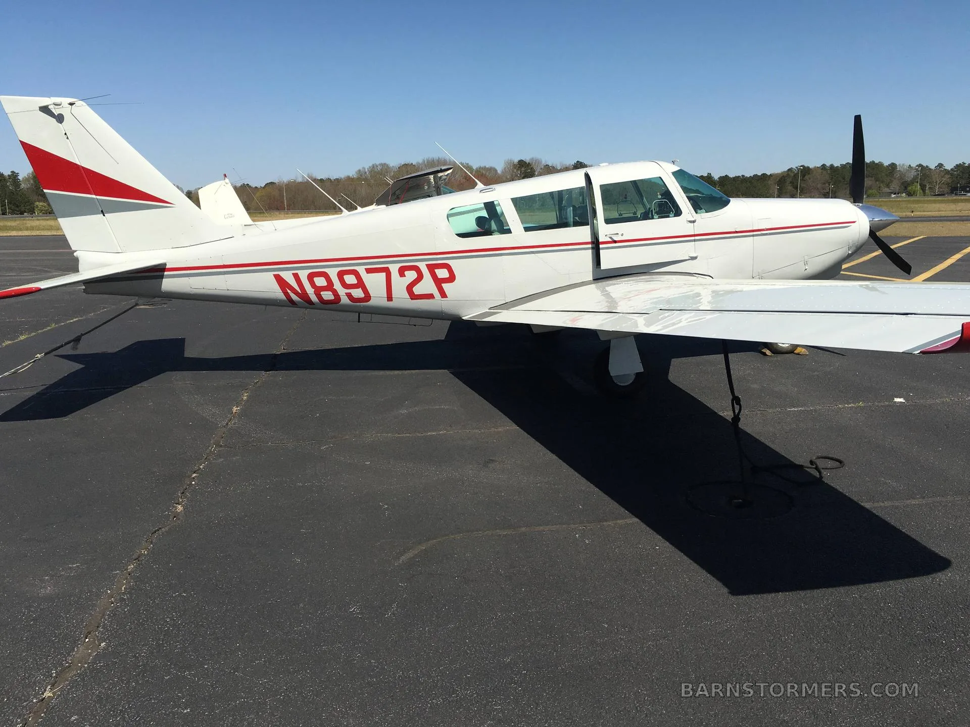 A white single-engine aircraft with red and grey stripes (N8972P) is parked on an asphalt ramp under a clear blue sky.