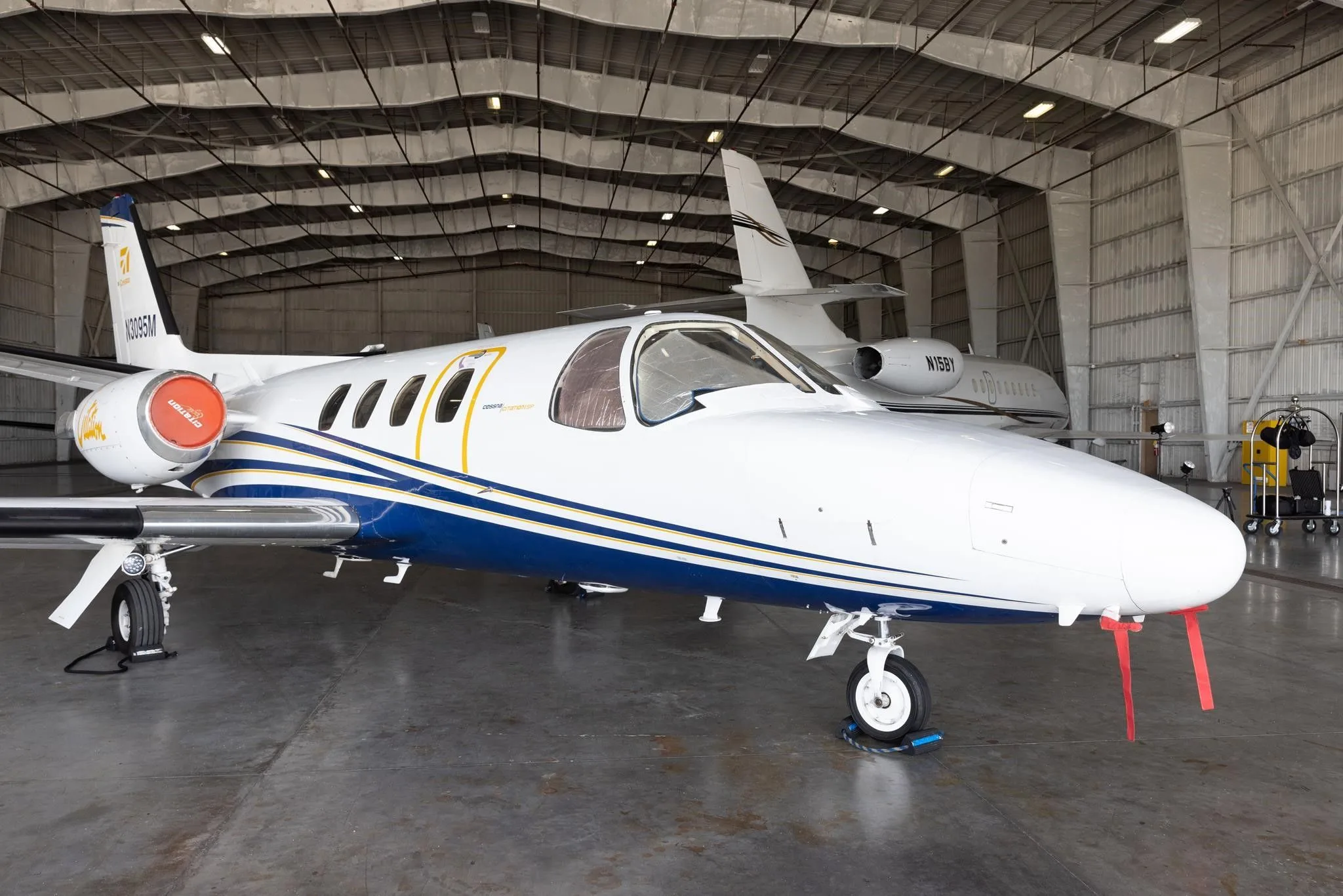 A front-side view of the white and blue Cessna Citation jet (N3095M) with red pitot tube covers, inside a hangar.