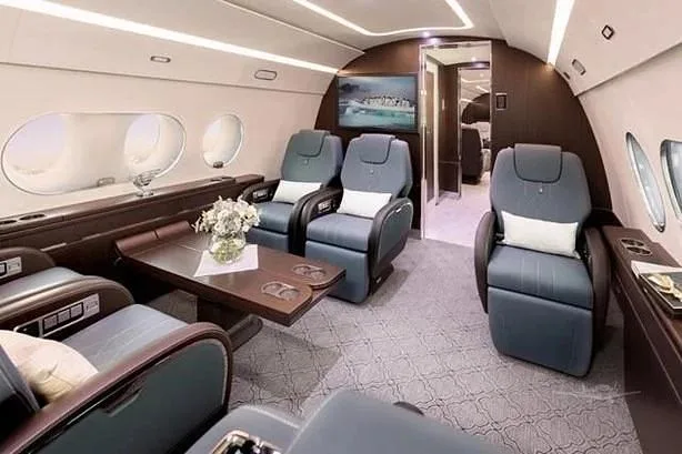 A luxurious aircraft cabin with blue leather seats, a dark wood table, and a large screen displaying a landscape, seen from a forward perspective.