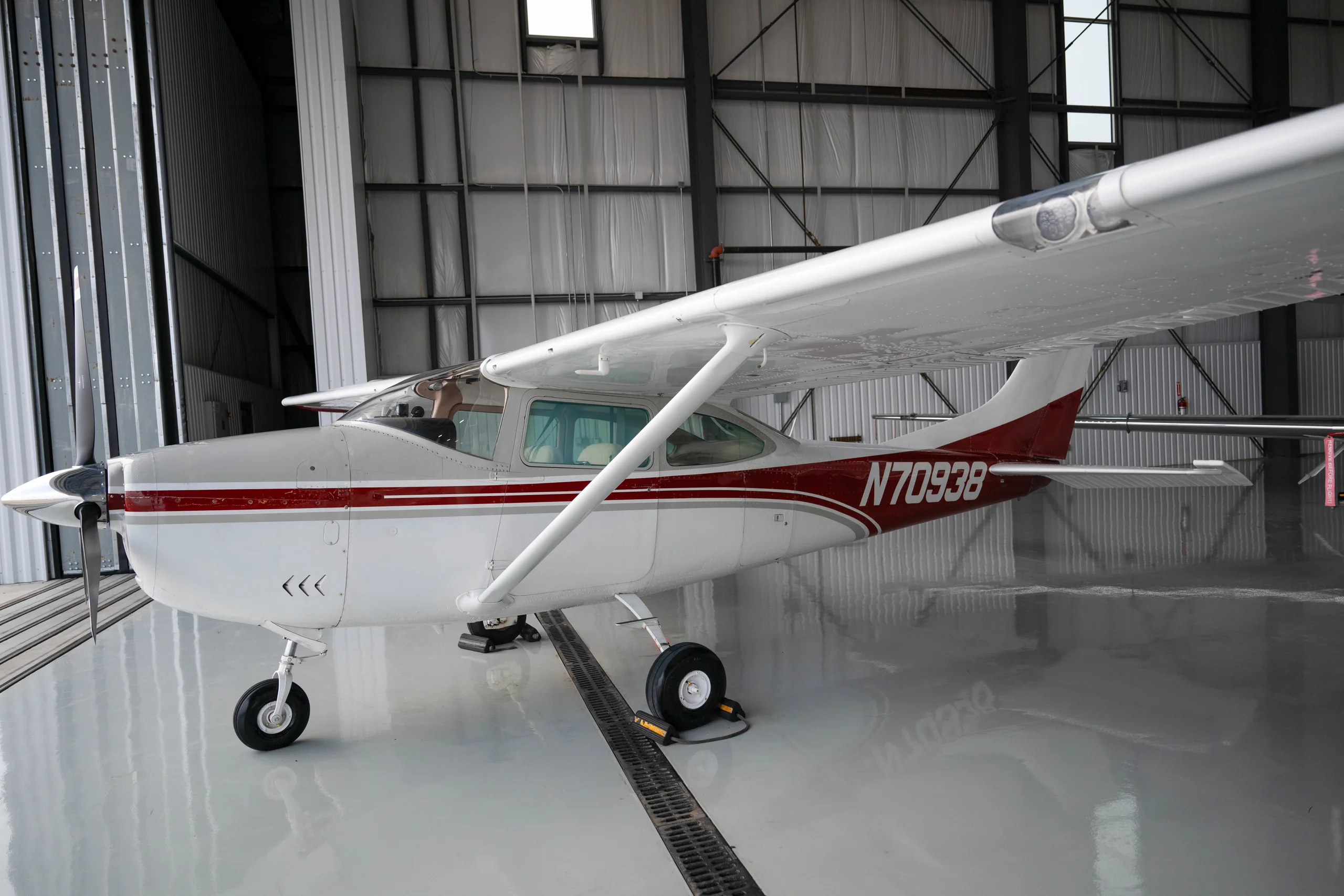 A white Cessna aircraft with red and gray stripes, registration N70938, parked in a hangar.