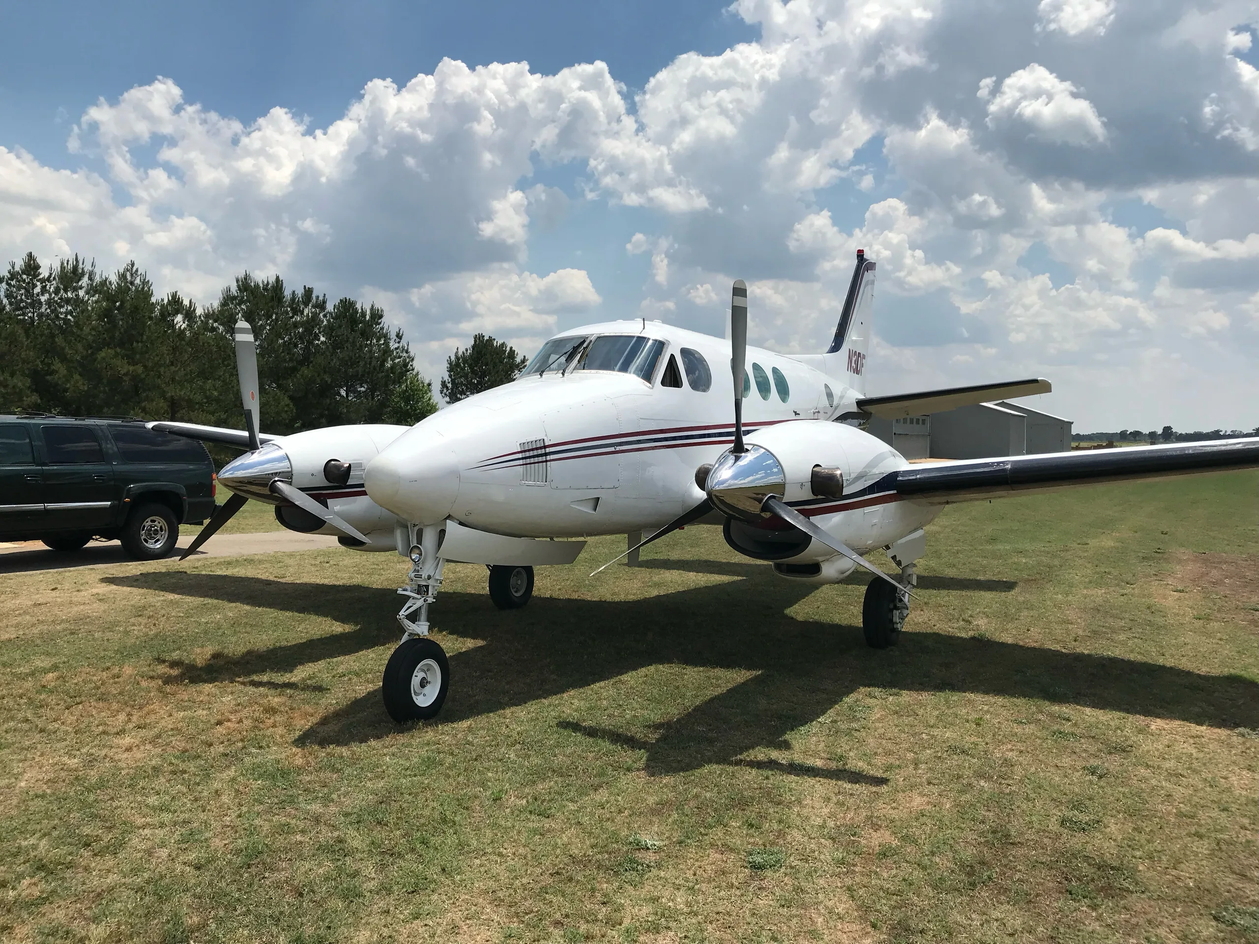 A white twin-engine turboprop aircraft with red and blue stripes is parked on a grassy field under a partly cloudy sky.