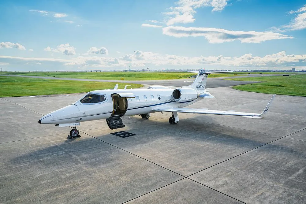 A white Learjet 45 business jet with blue and gold stripes, registration N90VJ, parked on an airport tarmac with its main door open under a partly cloudy sky.