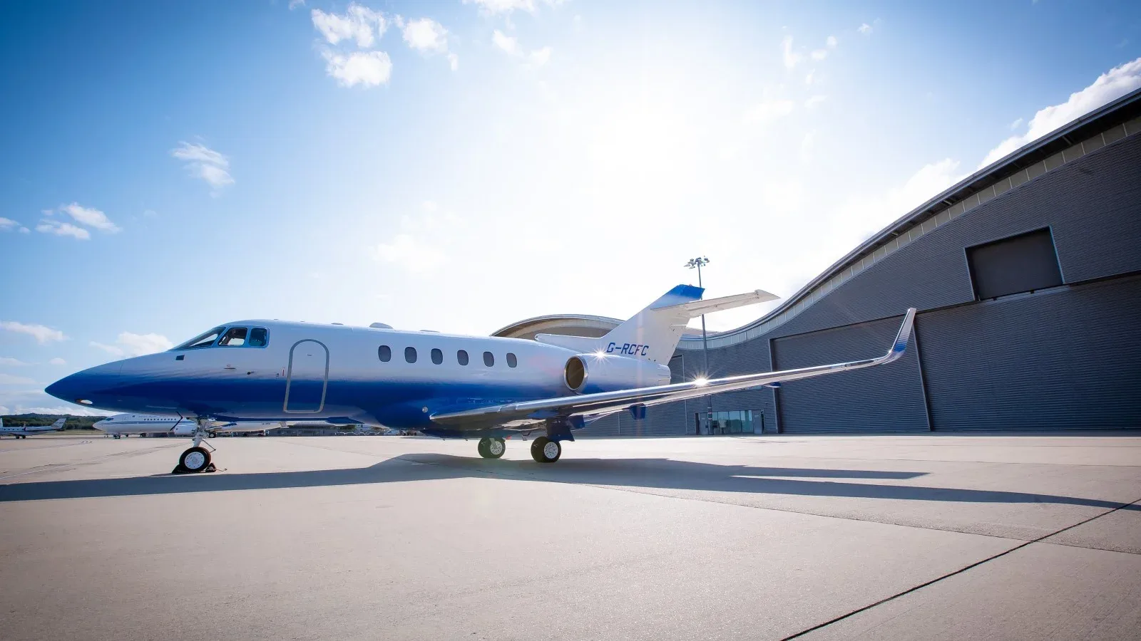 A blue and white jet sits on the tarmac in front of a hangar at Farnborough Airport.