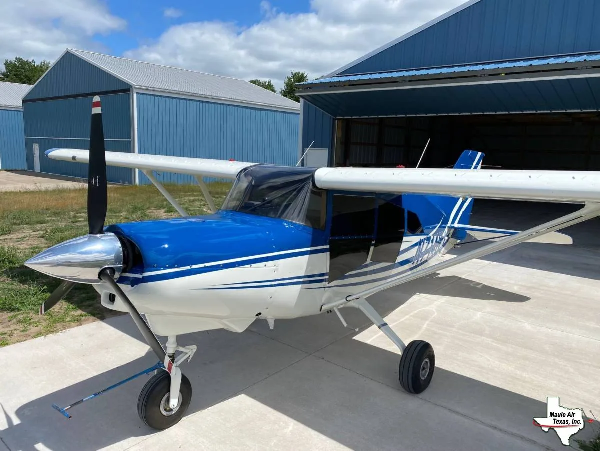 A blue and white single-engine propeller aircraft with a polished spinner is parked on concrete in front of a blue hangar under a partly cloudy sky.