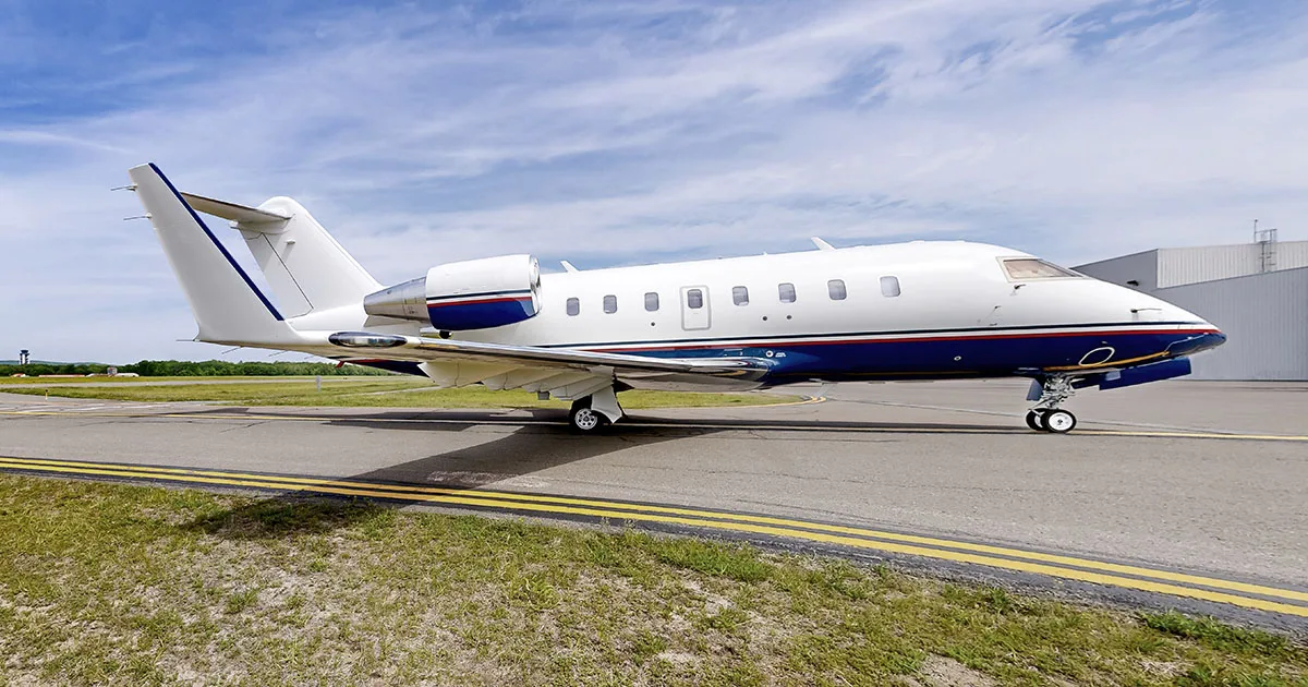 A white and blue Bombardier Challenger business jet is parked on a tarmac with a hangar in the background.