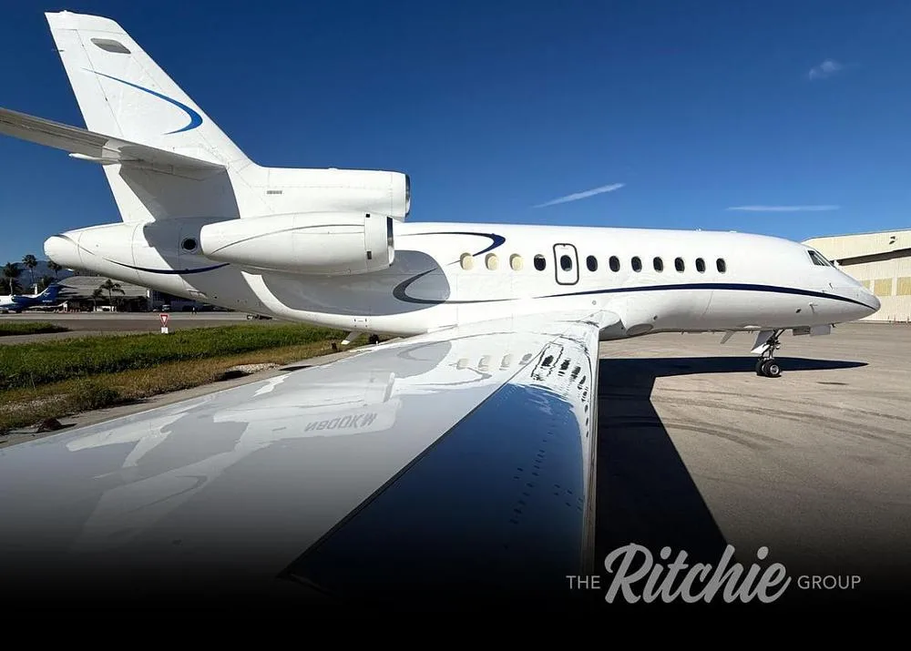A white Dassault Falcon business jet with blue accents, showing the tail and wing, parked on a tarmac under a clear blue sky.