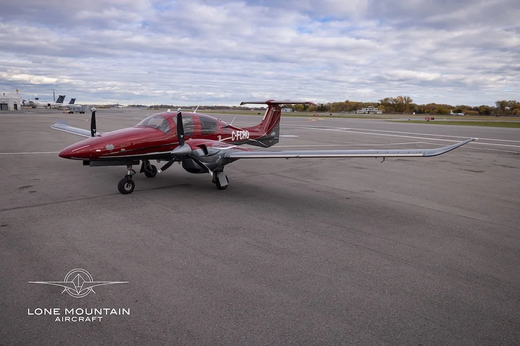 A red and grey Diamond DA62 aircraft with registration C-FCRQ parked on an airfield, viewed from the front.