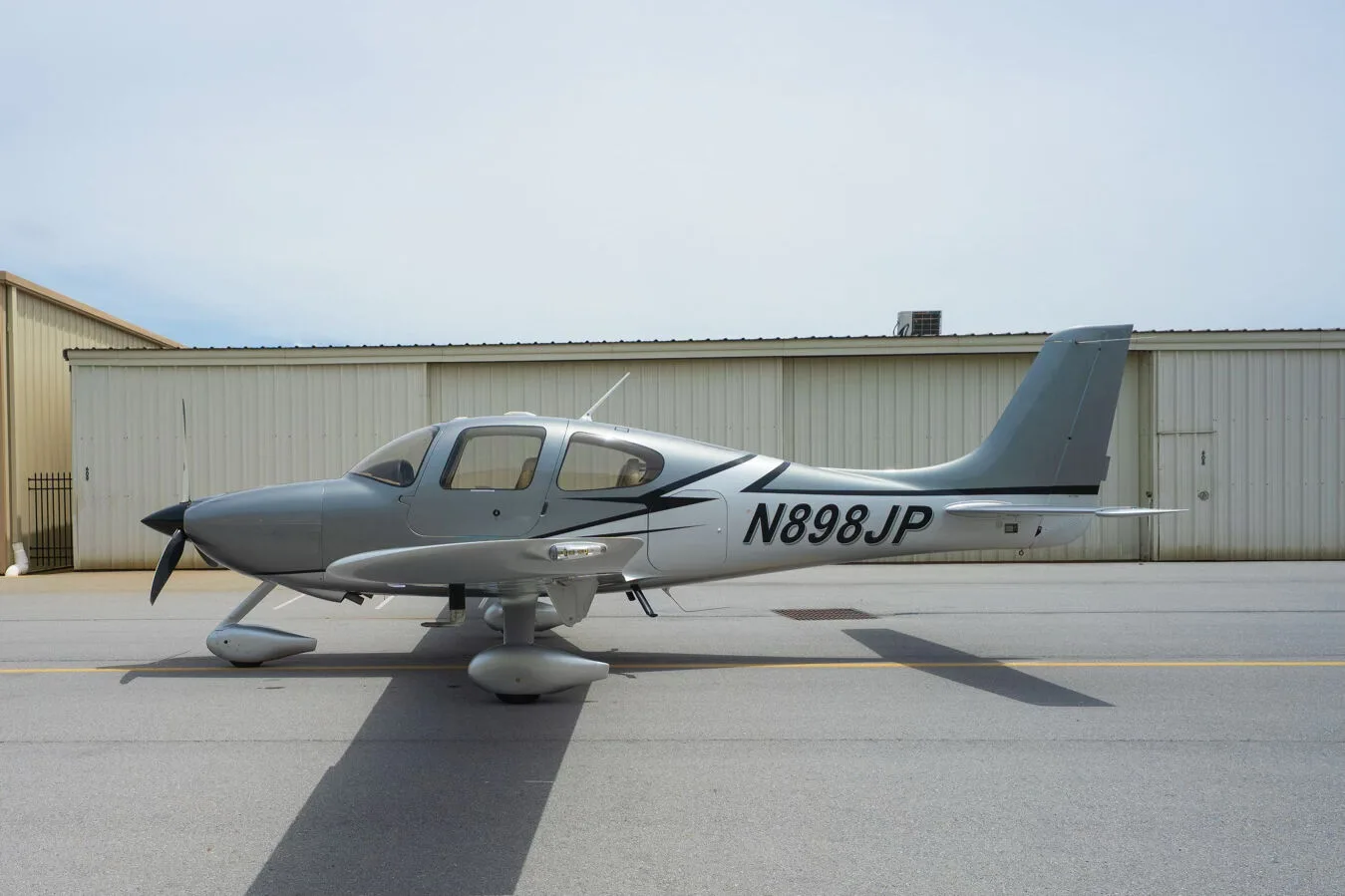 A silver Cirrus SR22 aircraft with black and white accents, registration N898JP, parked on an airfield next to hangars.