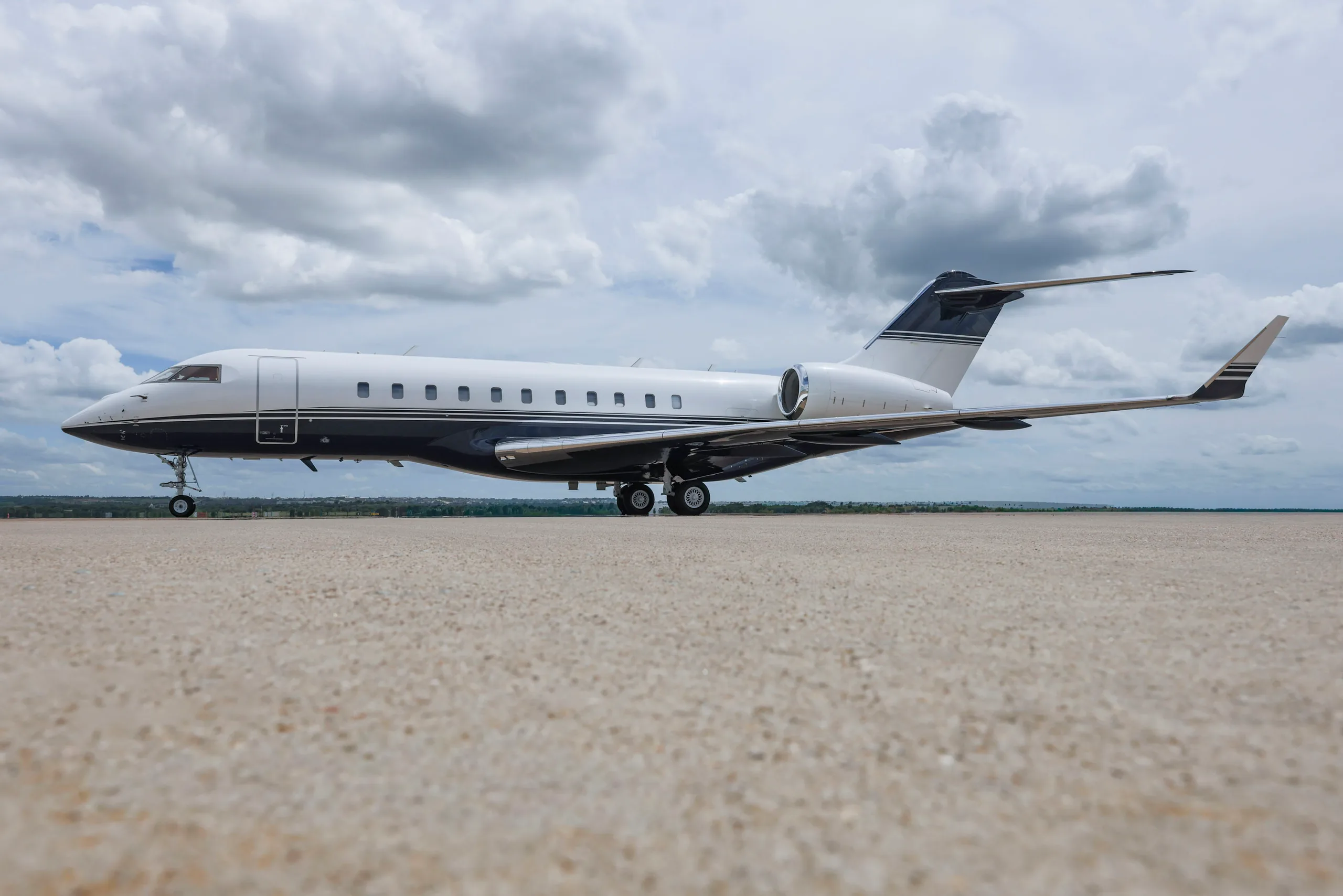 A white and dark blue Bombardier Global Express business jet is parked on an airfield under a cloudy sky.