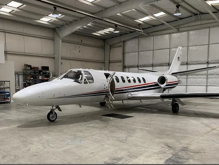 A white private jet with red and blue stripes is parked inside a large hangar with its main door open.