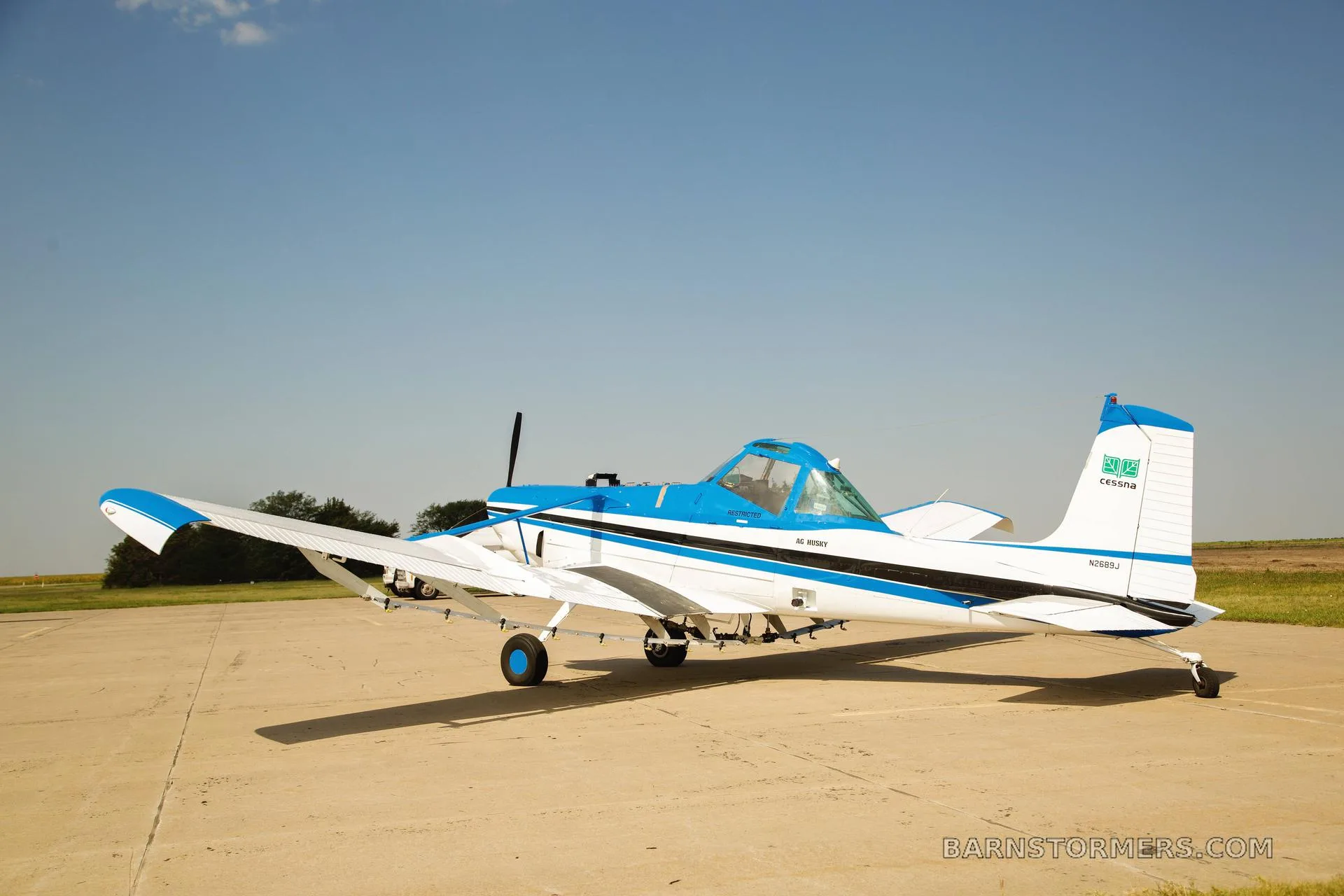 A white and blue agricultural aircraft, possibly a Cessna Ag Husky, is parked on a tarmac under a clear blue sky.
