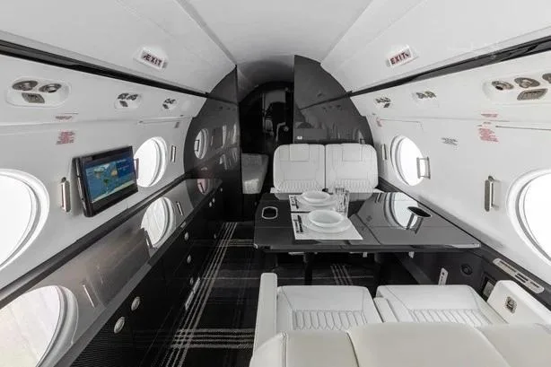 A private jet cabin interior with a dining table set for two, white seats, and a dark plaid carpet.
