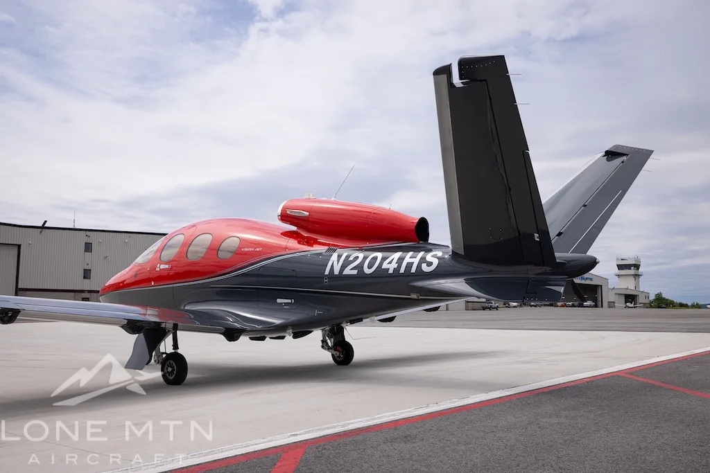 A rear-side view of the Cirrus Vision Jet N204HS in red and grey, highlighting the engine intake and V-tail, parked on an airfield.