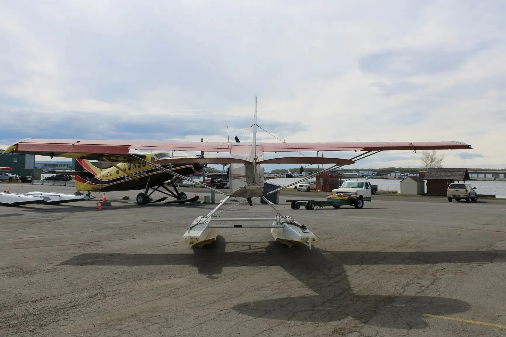 A rear view of the red and grey floatplane, showing its tail and floats, parked on an apron with other aircraft.
