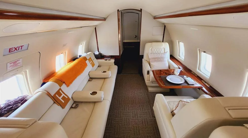 A wide view of the private jet's cream-colored cabin interior with facing seats, a table, and an "EXIT" sign.