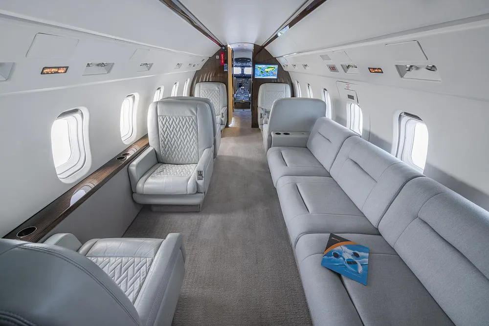 The spacious interior cabin of a business jet features light gray individual seats and a long sofa, looking towards the cockpit.