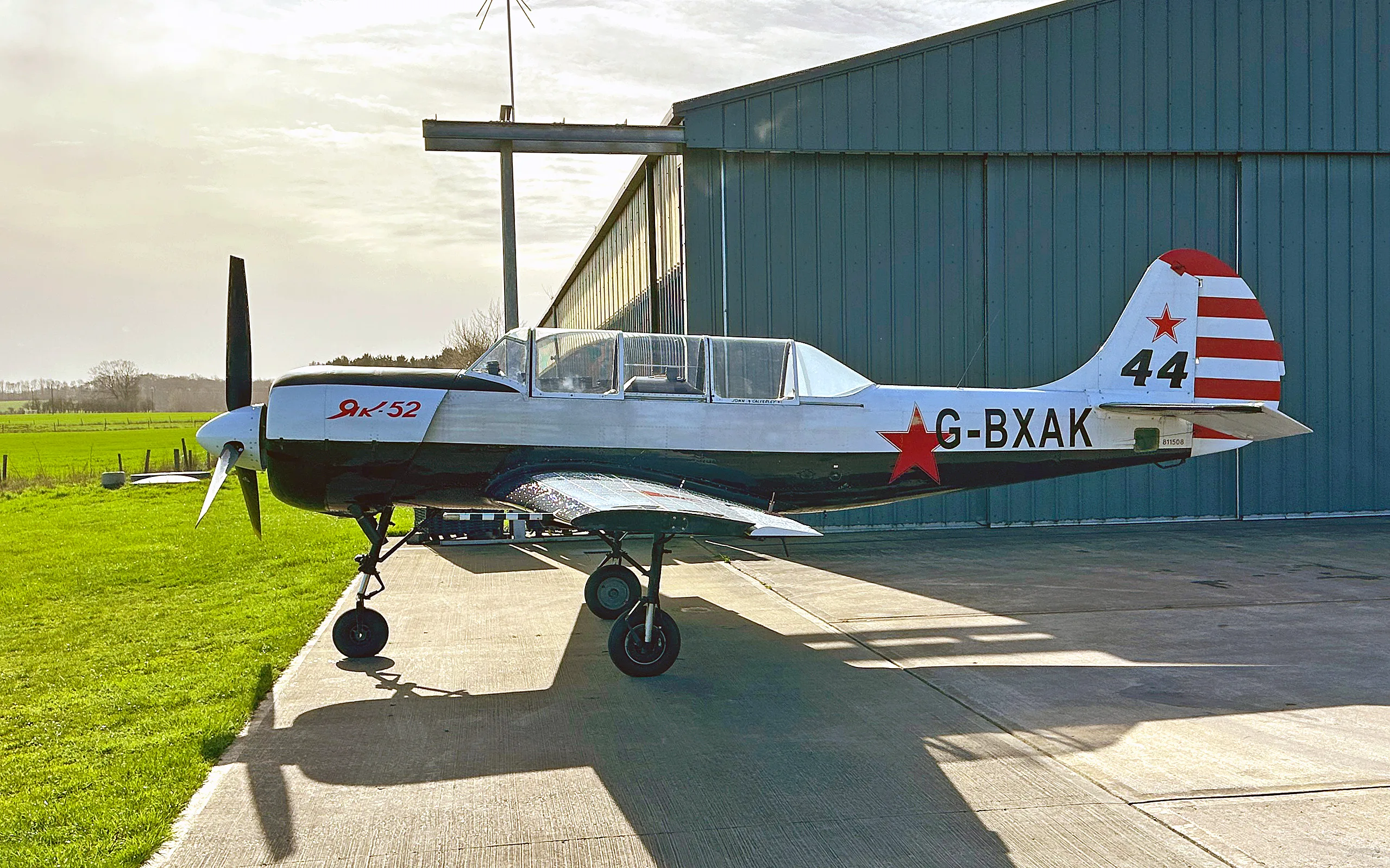 A white and black Yak-52 aircraft with red stars and tail stripes, parked on a concrete pad in front of a hangar under a sunny sky.