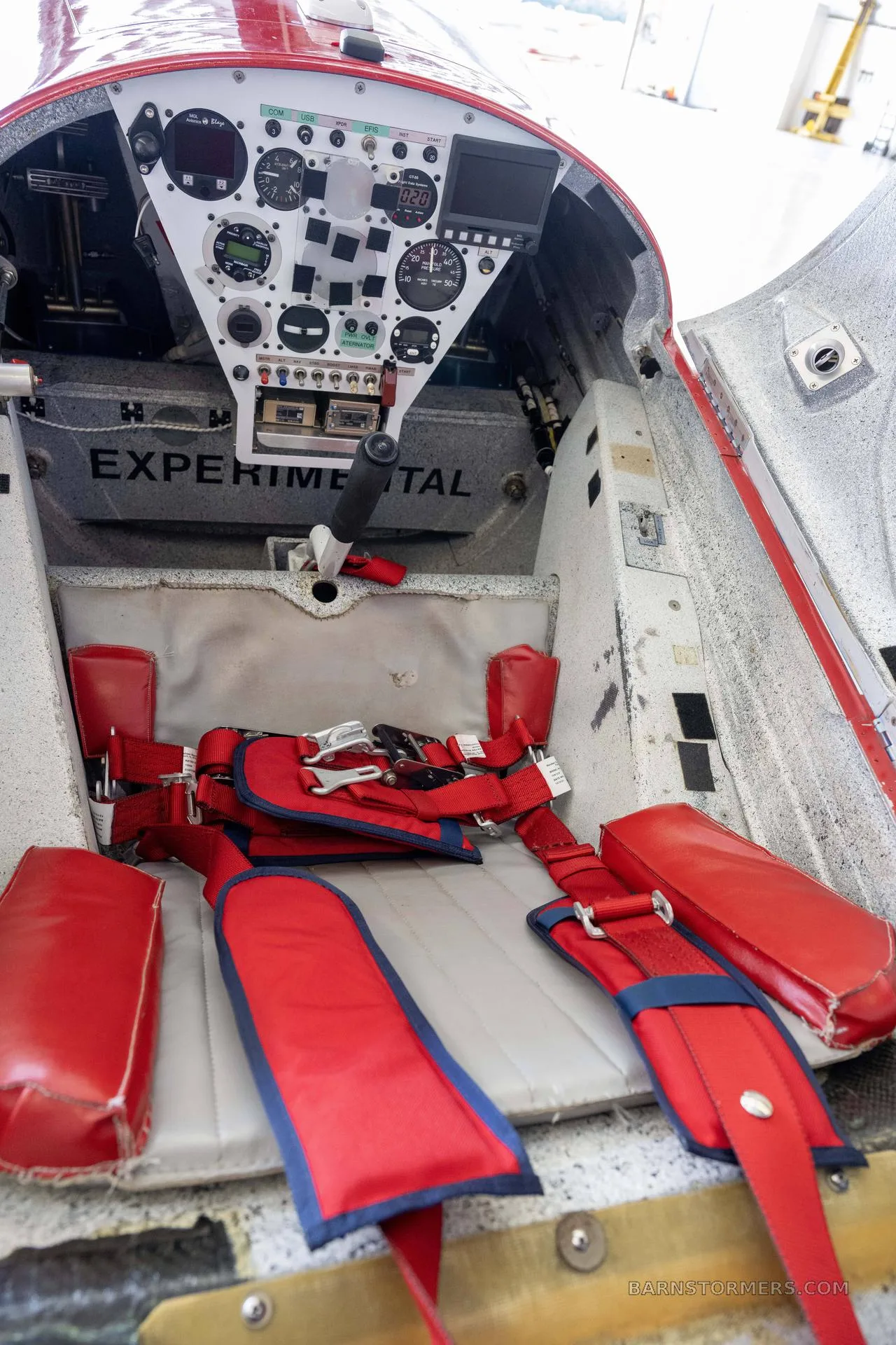 The open cockpit of an experimental aircraft, showing the instrument panel, control stick, and red safety harness.