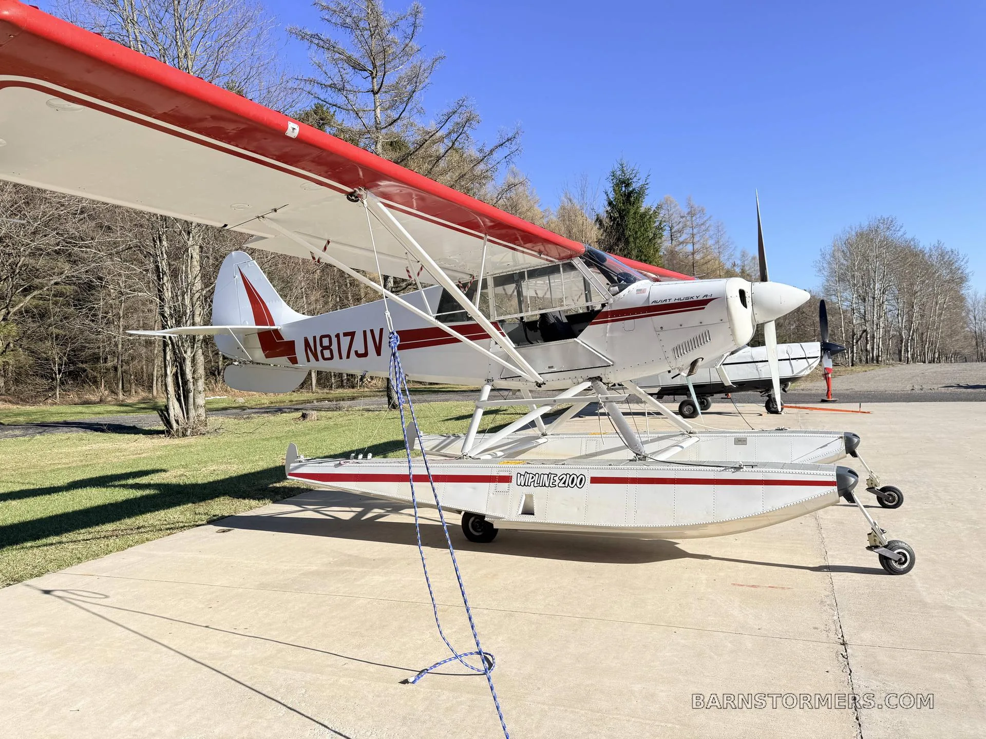 A white and red Aviat Husky A-1 floatplane (N817JV) parked on a concrete pad with trees in the background.