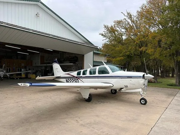A white Beechcraft Bonanza with blue and dark stripes, registration N222SN, parked on a concrete pad outside a hangar with trees in the background.