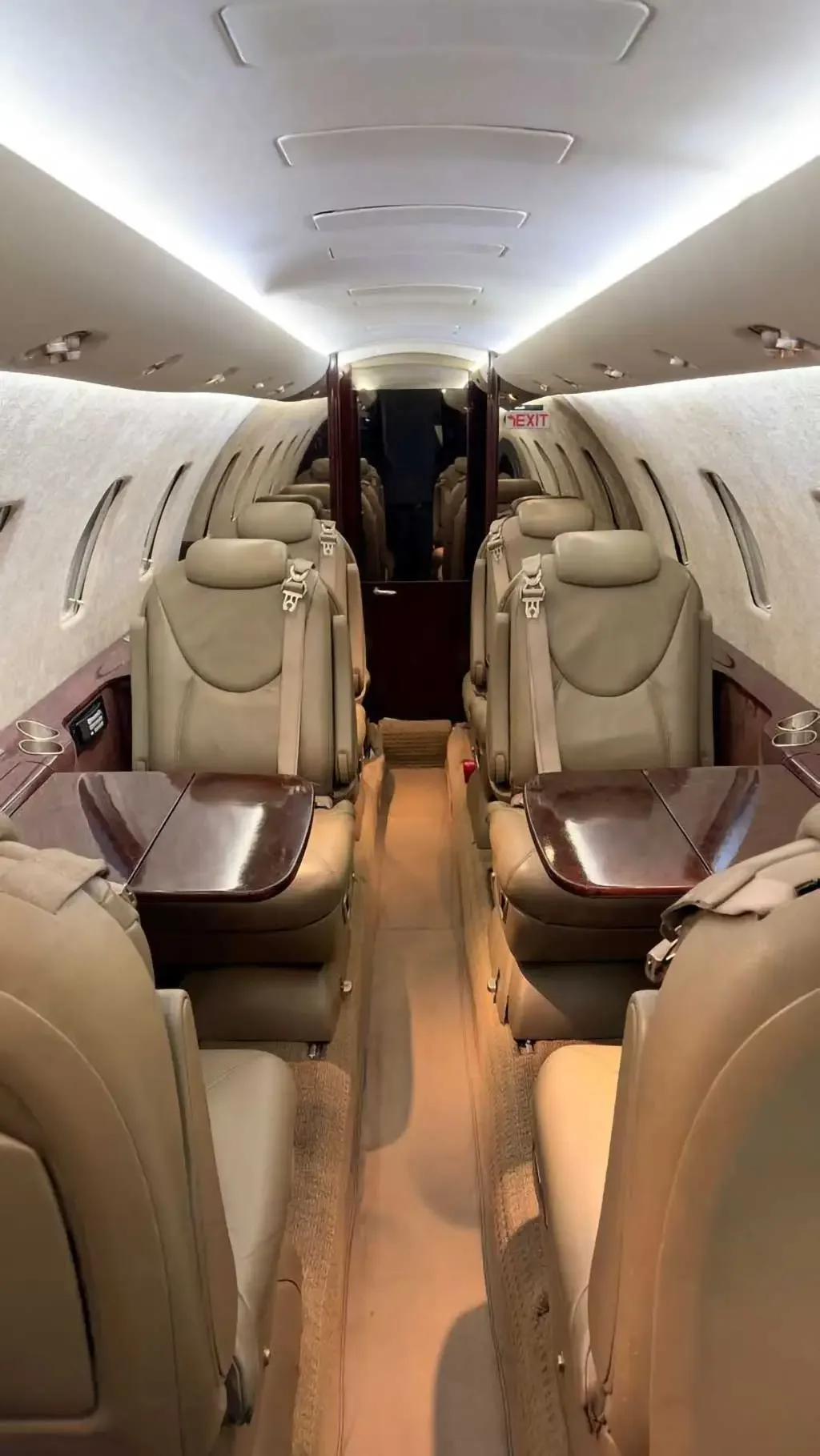 View of the aircraft cabin interior with beige leather seats and wooden tables, looking towards the rear of the cabin.