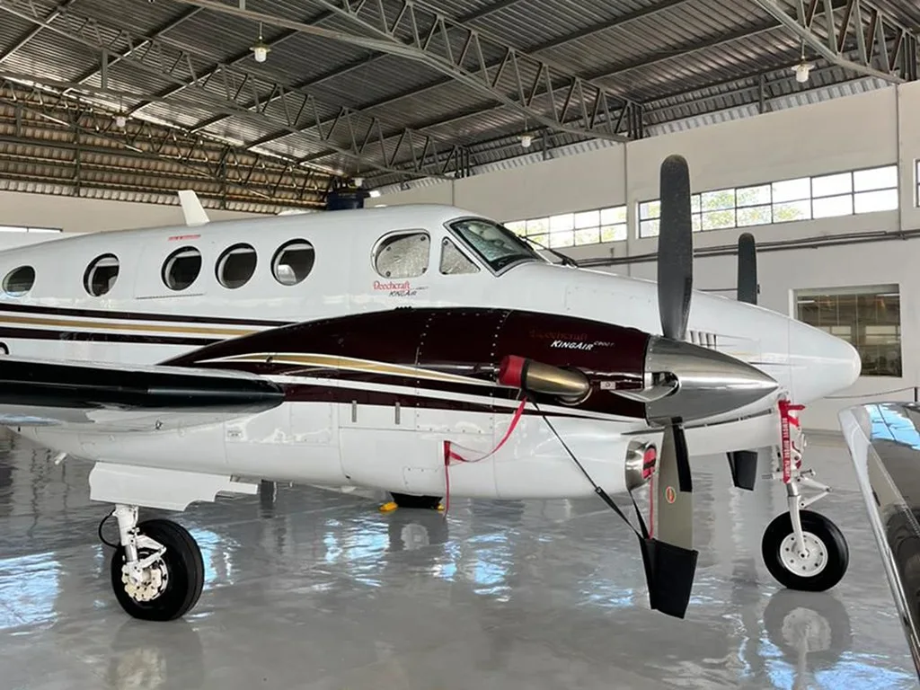Side view of the front section of a white and maroon Beechcraft King Air 350, showing the propeller and cockpit windows, inside a hangar.