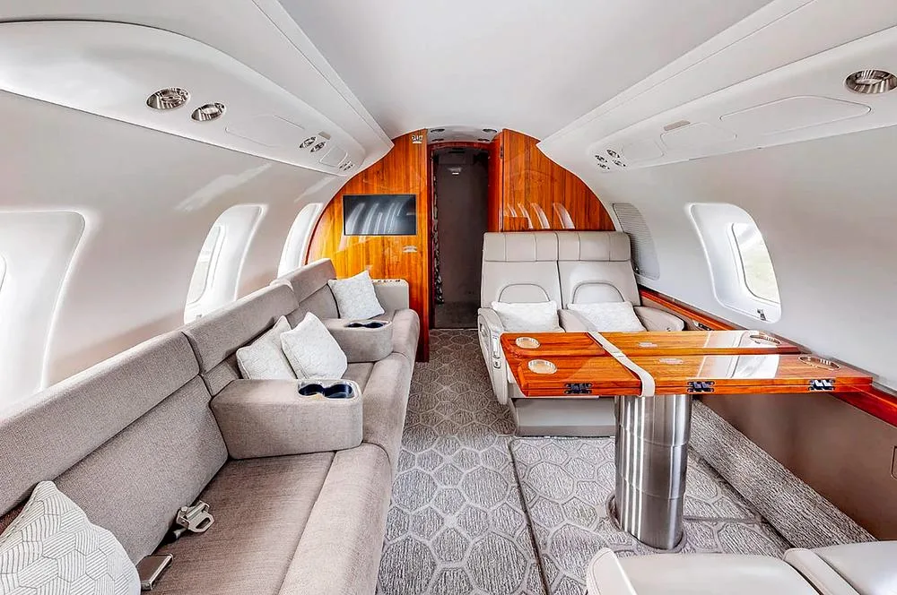 The spacious aircraft cabin includes a grey sofa, a polished wooden table, and light-colored seating.