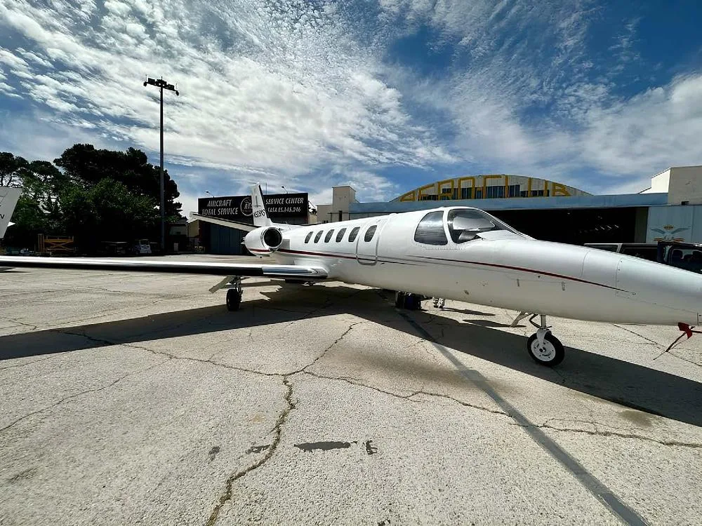 A white Cessna CitationJet aircraft with a red stripe is parked on an airport tarmac, viewed from the side, under a cloudy sky.