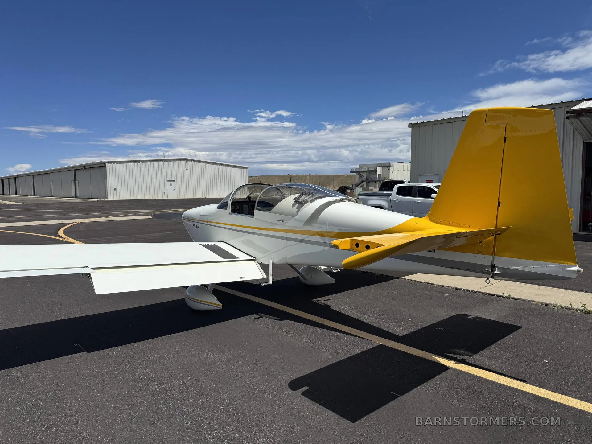 A rear-side view of a white RV-9A aircraft with a prominent yellow tail, parked on an asphalt ramp under a blue sky.