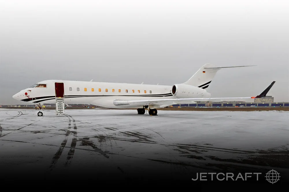 A white Bombardier Global Express business jet with black and gold stripes on a snowy tarmac, with its main door open and stairs extended.
