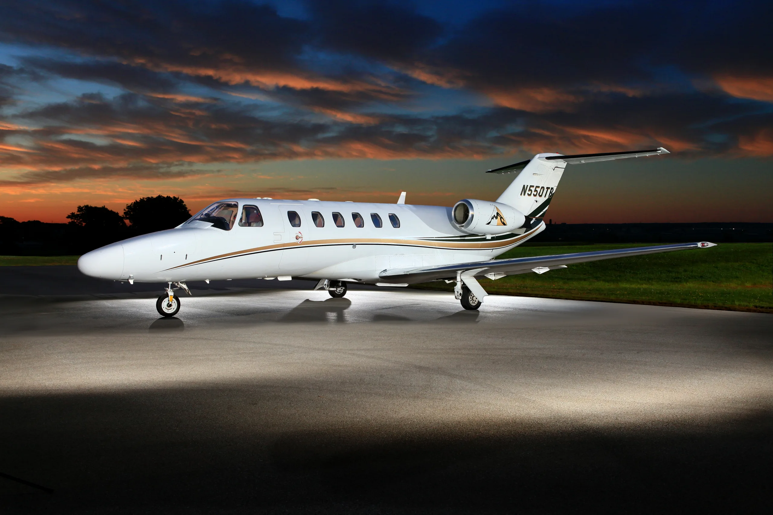 A white Cessna Citation V (N550TB) with gold and dark green stripes is parked on a tarmac at sunset, illuminated by ground lights.