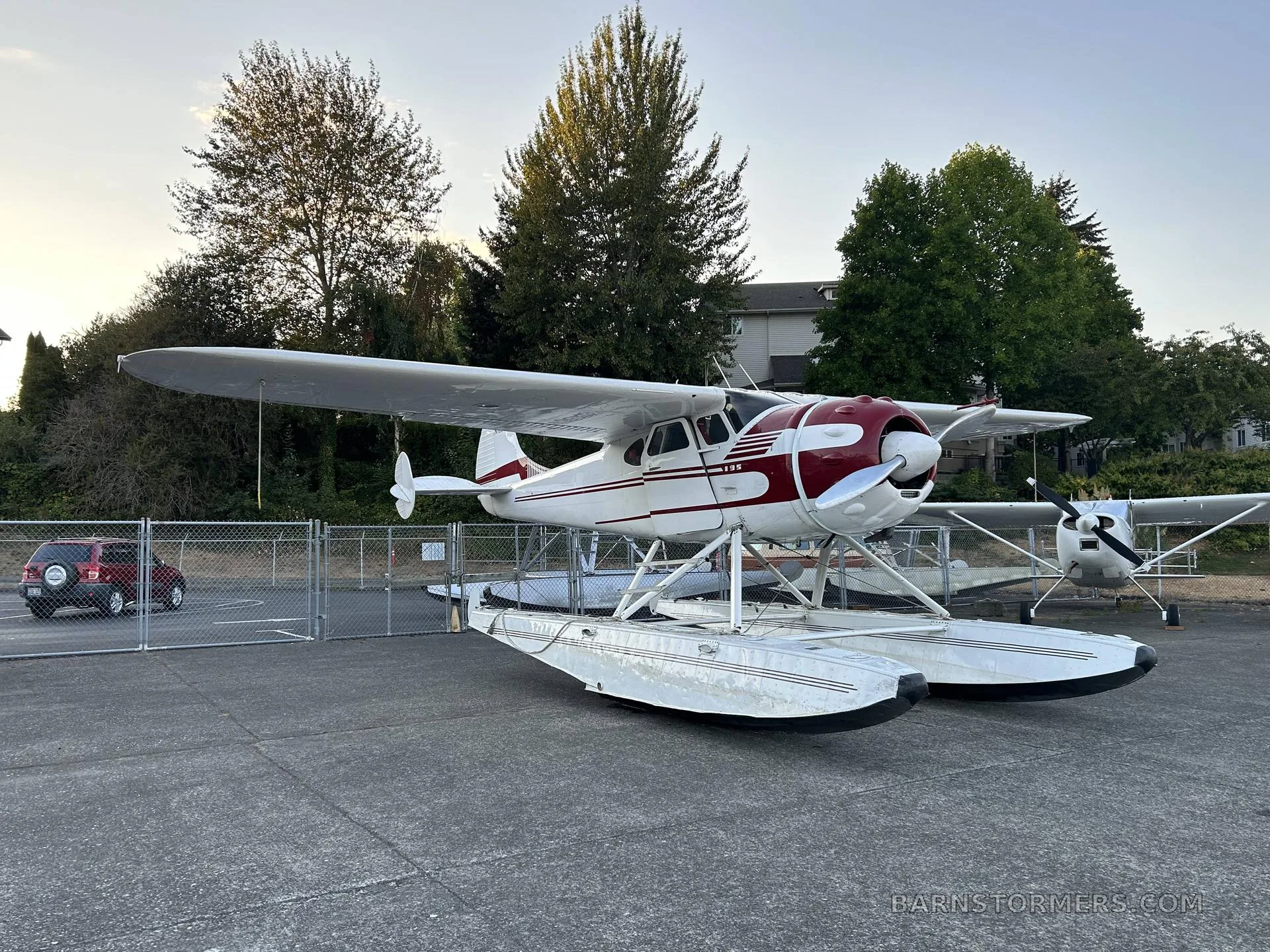 A white and red floatplane, likely a Cessna 180 or 185, parked on concrete next to a fence with trees and a building in the background.