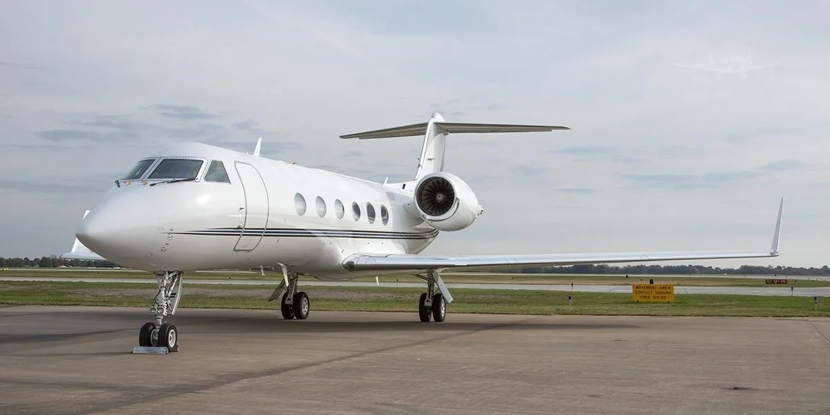 A white Gulfstream business jet with a thin dark stripe is parked on an airport tarmac under a cloudy sky.
