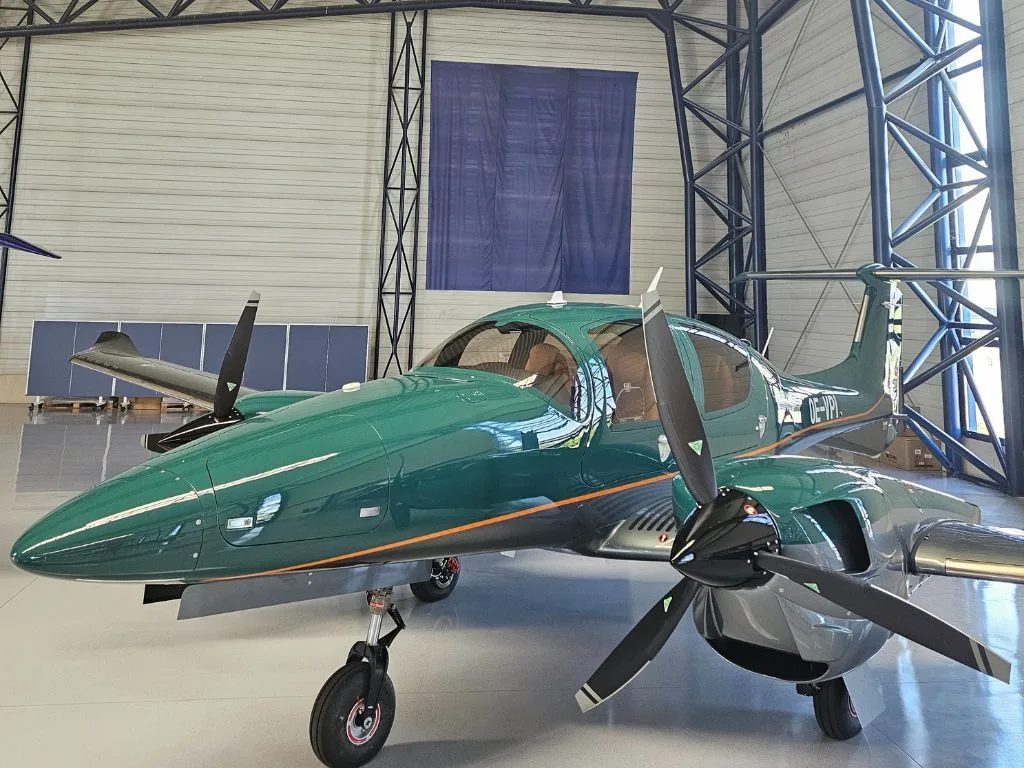 A dark green Diamond DA62 aircraft with an orange stripe is parked inside a hangar.